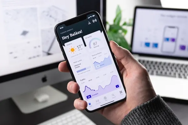 Hand holding smartphone with analytics dashboard and laptop in background